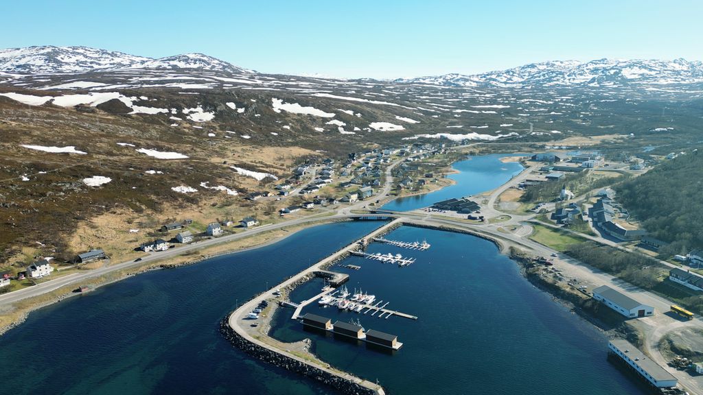 Lite tettsted ved en fjord. Molo og småbåthavn i forgrunnen. Fjell med snø i bakgrunnen. Foto.