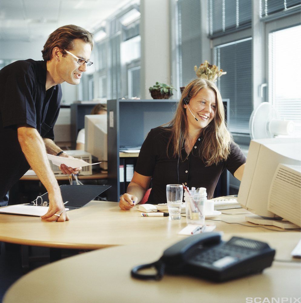 To personer i et kontorlandskap, hvor den ene har hodetelefoner og ser på en dataskjerm. Foto.