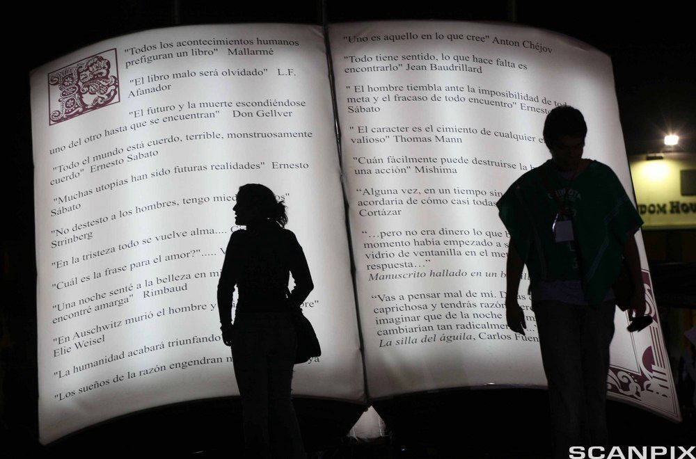 Silhuetter av to figurer mot en skulptur utforma som ei bok. Foto. 