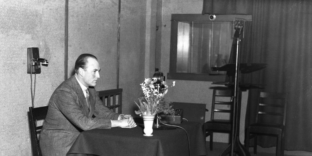 Daværende kronprins Olav sitter ved et bord i et radiostudio. Foto.