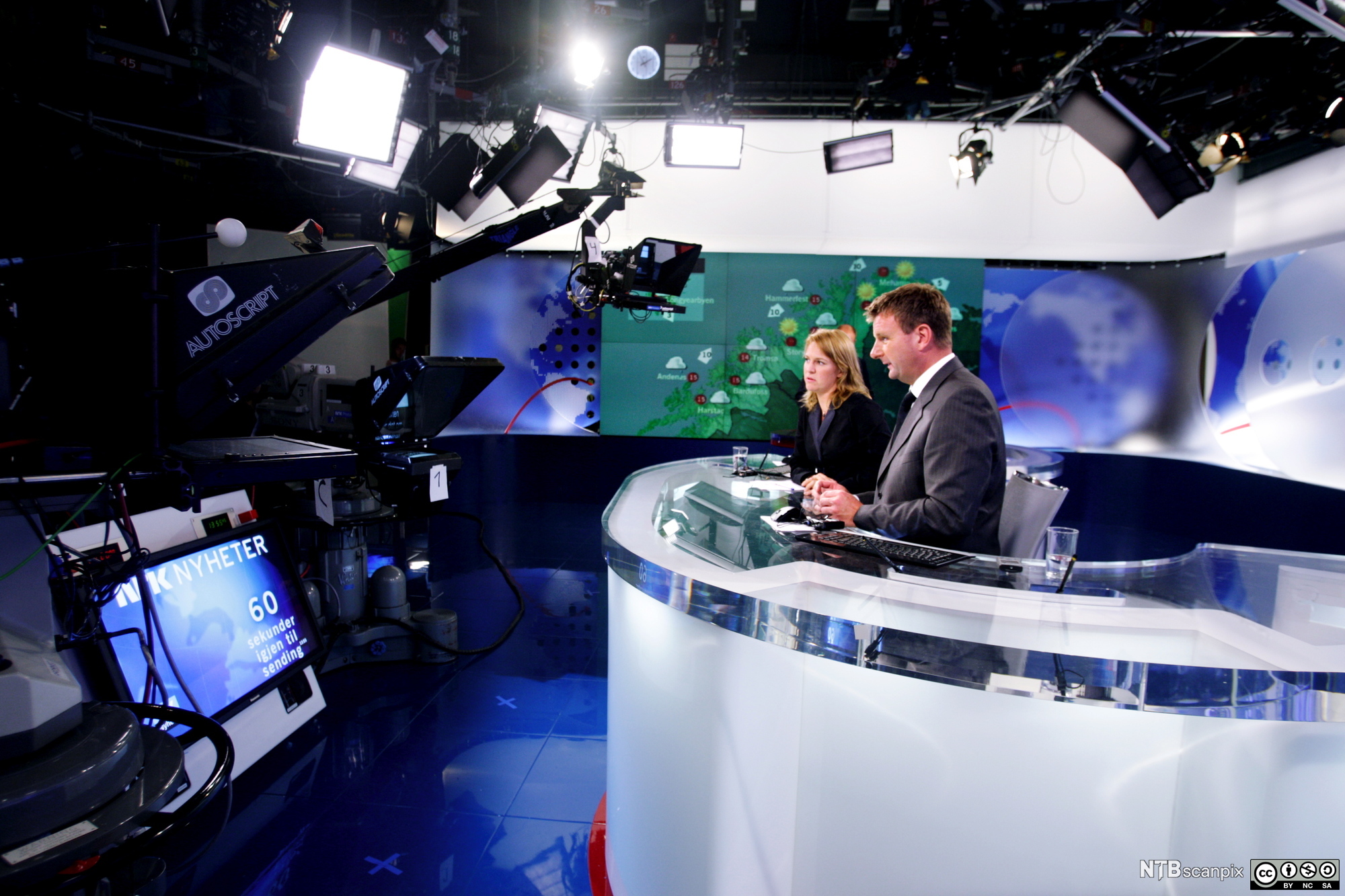 Foto av programlederne Gry Blekastad Almås og Jon Gelius i NRKs nyhetsstudio. I bildet er det flere kameraer og lys og en skjerm med info om direktesending, og i bakgrunnen ser vi værkartet i en annen del av tv-studioet.