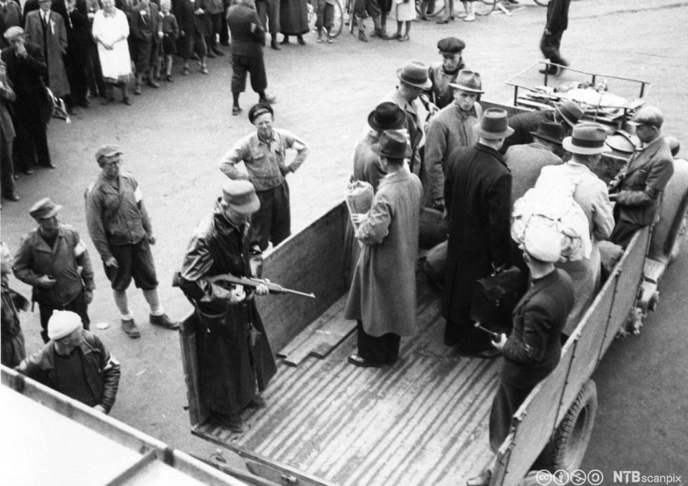 Noen menn står på lasteplanet av en lastebil. Det står også to menn med gevær i enden av lasteplanet. Rundt står det mange mennesker. Svart-hvitt-foto. 