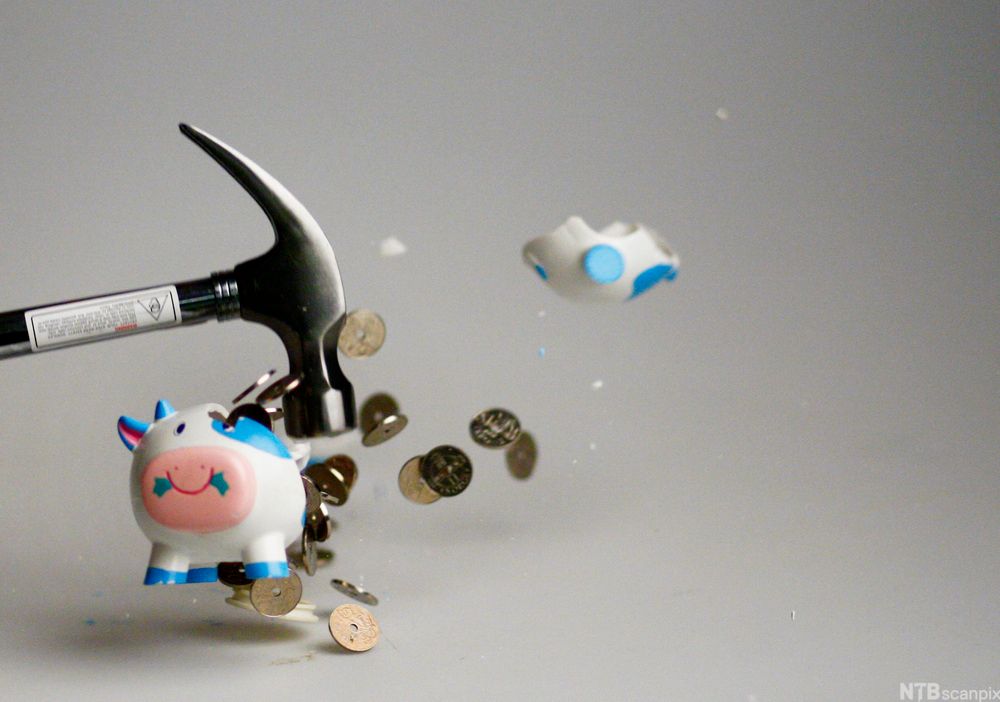 Foto av ein sparegris i porselen som blir slått sund med ein hammar. Myntar og bitar av sparegrisen fyk utover.