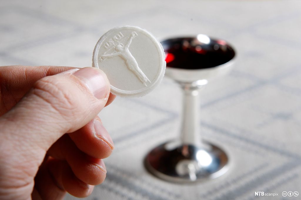 En hånd holder en rund, hvit skive. Bak et lite beger med rød vin. Foto.