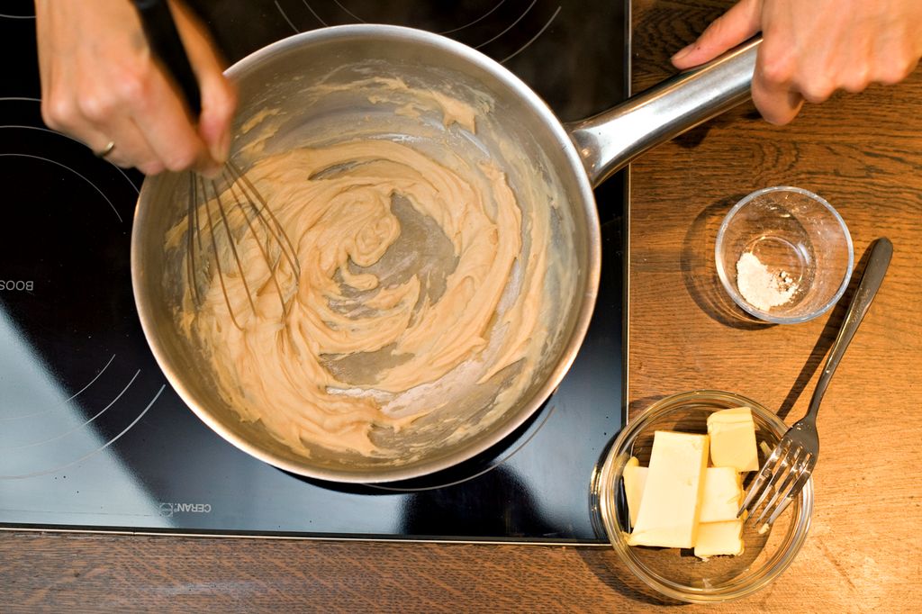 En hånd rører med en visp i ei panne med brun jevning. Foto.