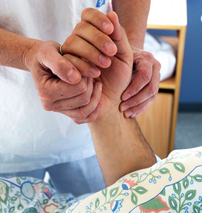 Helsefagarbeidar tek puls på ein pasient som ligg i ei seng. Foto.