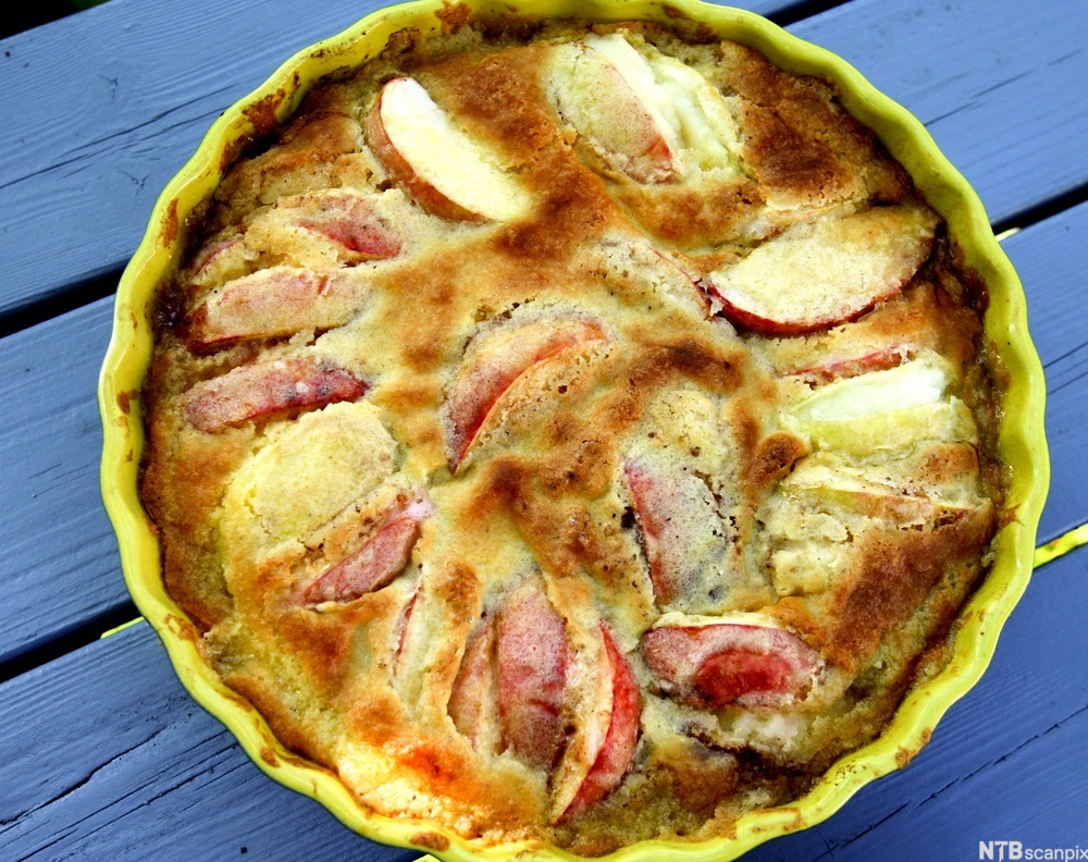 Eplekake i rund keramikkform. Innholdet har en sprøstekt og ujevn overflate. Mange biter av røde epler stikker opp av deigen. Foto.