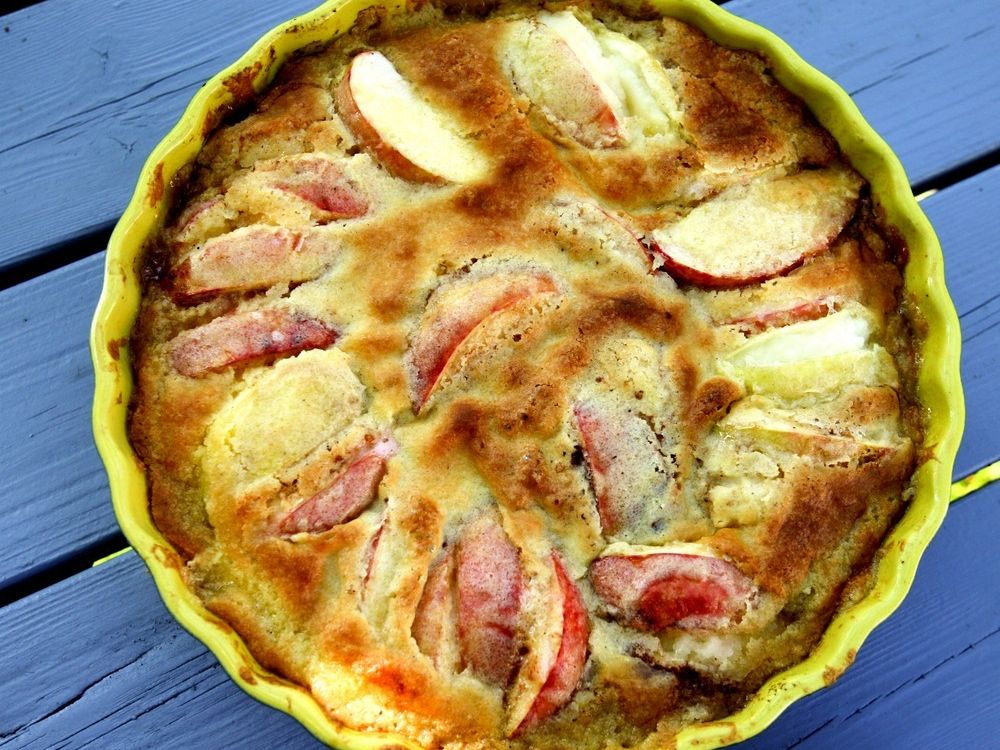 Eplekake i rund keramikkform. Innholdet har en sprøstekt og ujevn overflate. Mange biter av røde epler stikker opp av deigen. Foto.