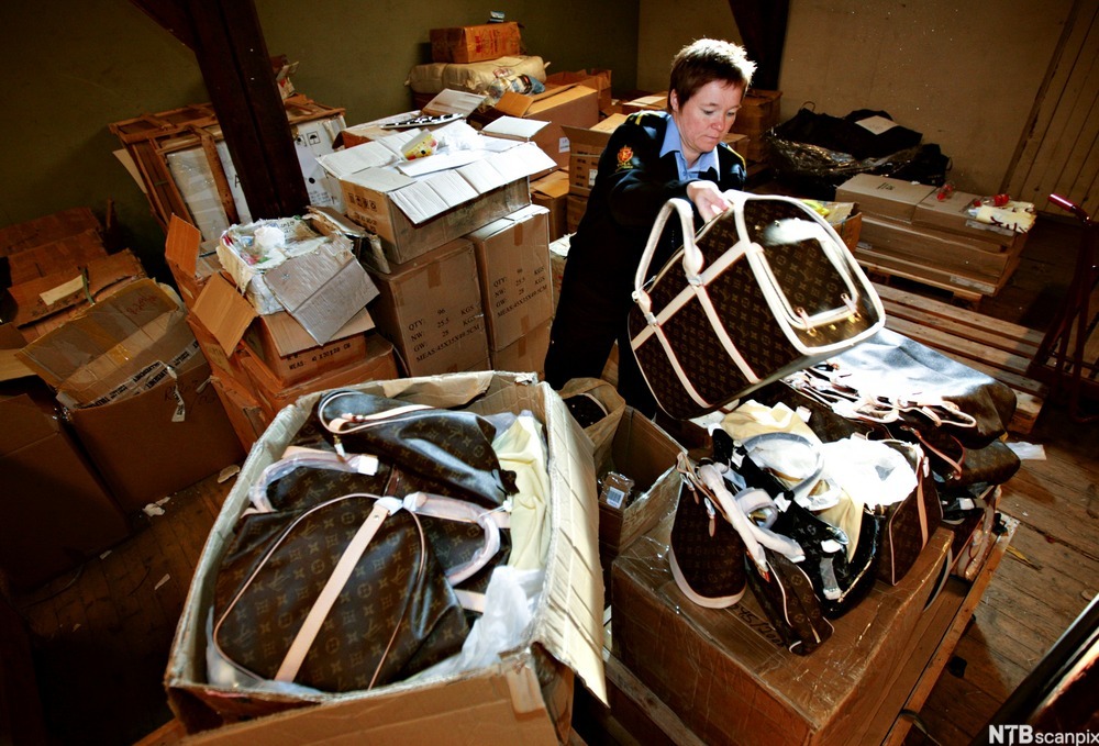 En tollbetjent på et rotet lagerrom pakker ut vesker med mønster som likner på det som produsenten Louis Vuitton er kjent for. Foto.
