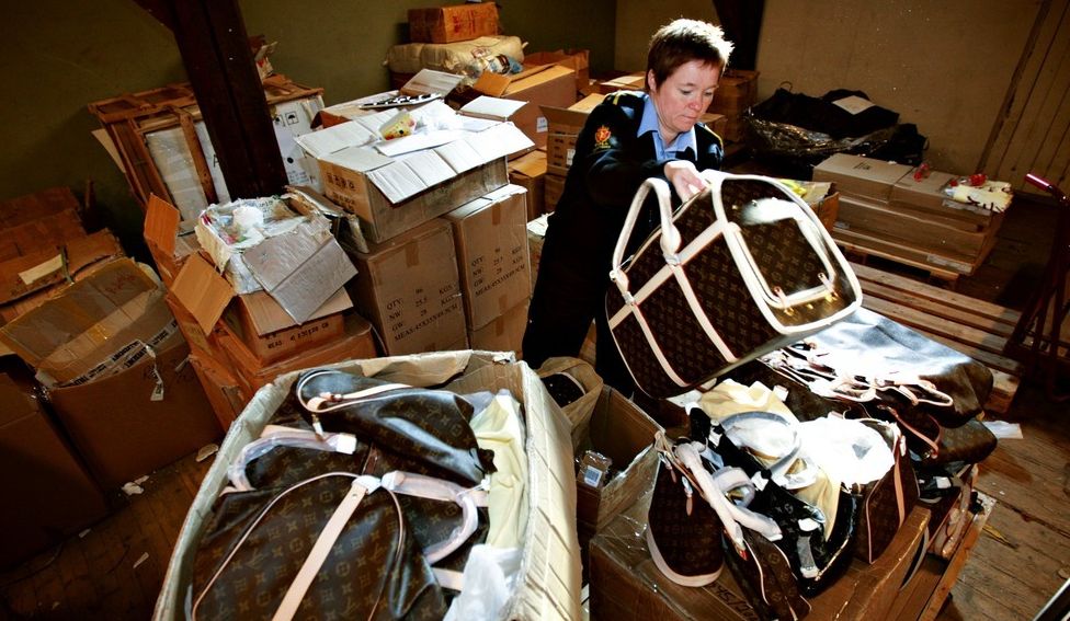 En tollbetjent på et rotet lagerrom pakker ut vesker med mønster som likner på det som produsenten Louis Vuitton er kjent for. Foto.