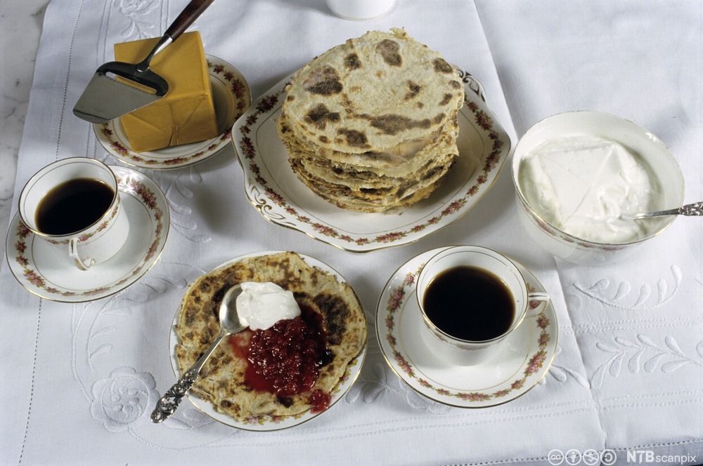 Et bord med hvit duk med et fat med lomper, brunost, rømme og to kaffekopper.  Foto.