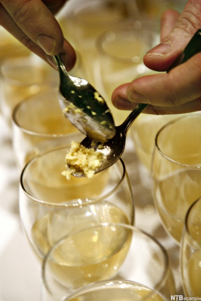 En person skraper revet ingefær fra en skje ned i et glass. Foto.