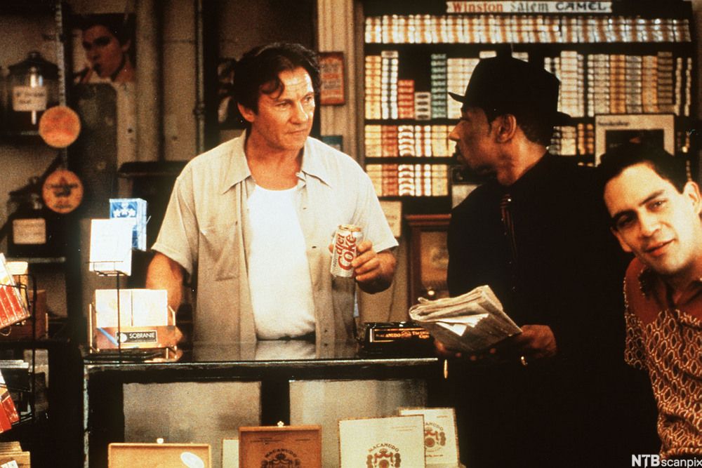 A scene from the film Smoke. Auggie Wren is standing behind the counter of his shop. Two other customers are in the picture. Photo.
