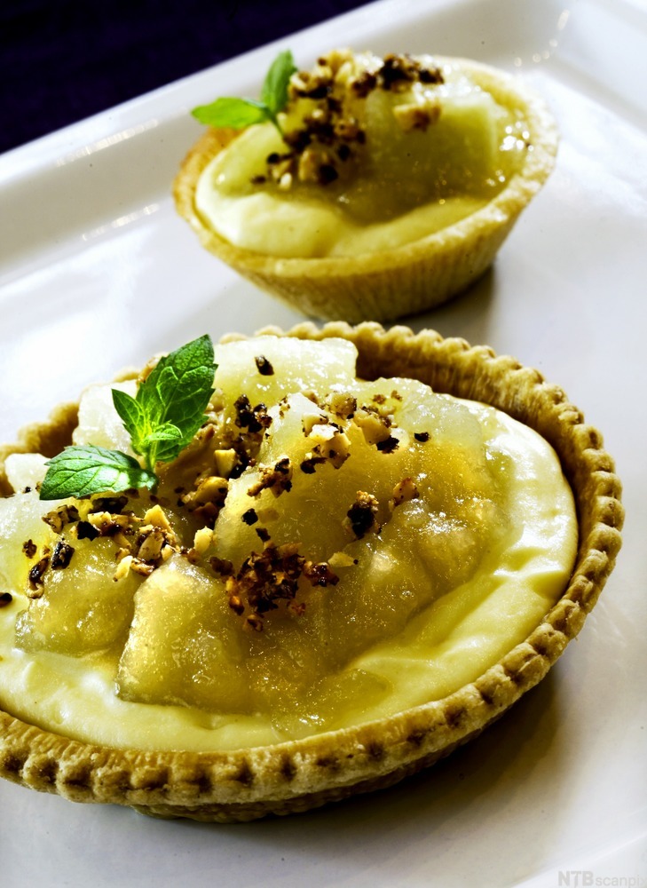 Liten dessertskål av butterdeig fylt med en gul, kremaktig masse. Et grønt blad og små kornaktige biter er strødd på toppen. Foto.