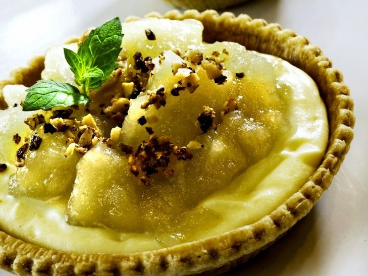 Liten dessertskål av butterdeig fylt med en gul, kremaktig masse. Et grønt blad og små kornaktige biter er strødd på toppen. Foto.