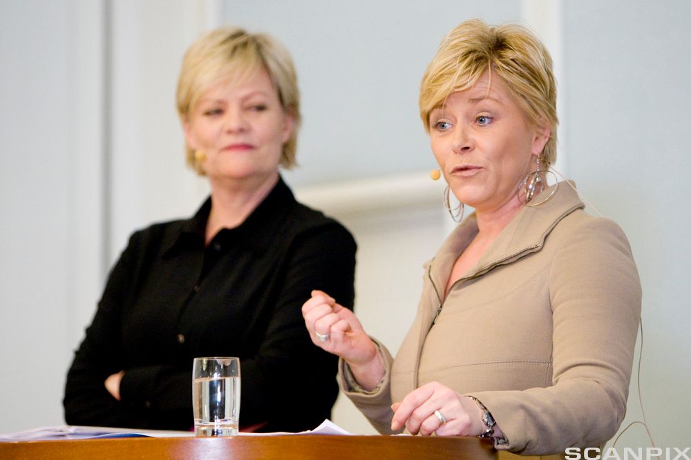 Tidligere Frp-leder Siv Jensen står bak et bord og gestikulerer i en debatt. Ved siden av henne står tidligere kunnskaps- og finansminister for SV, Kristin Halvorsen, med armene i kors og ser på Jensen. Begge har myggmikrofon på kinnet. Foto.