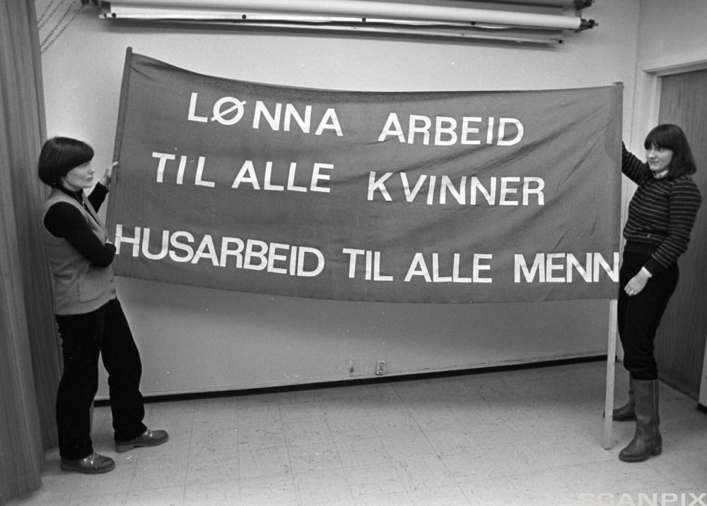 To kvinner som held ei fane med teksten "lønna arbeid til alle kvinner, husarbeid til alle menn". Svart-kvitt-foto.