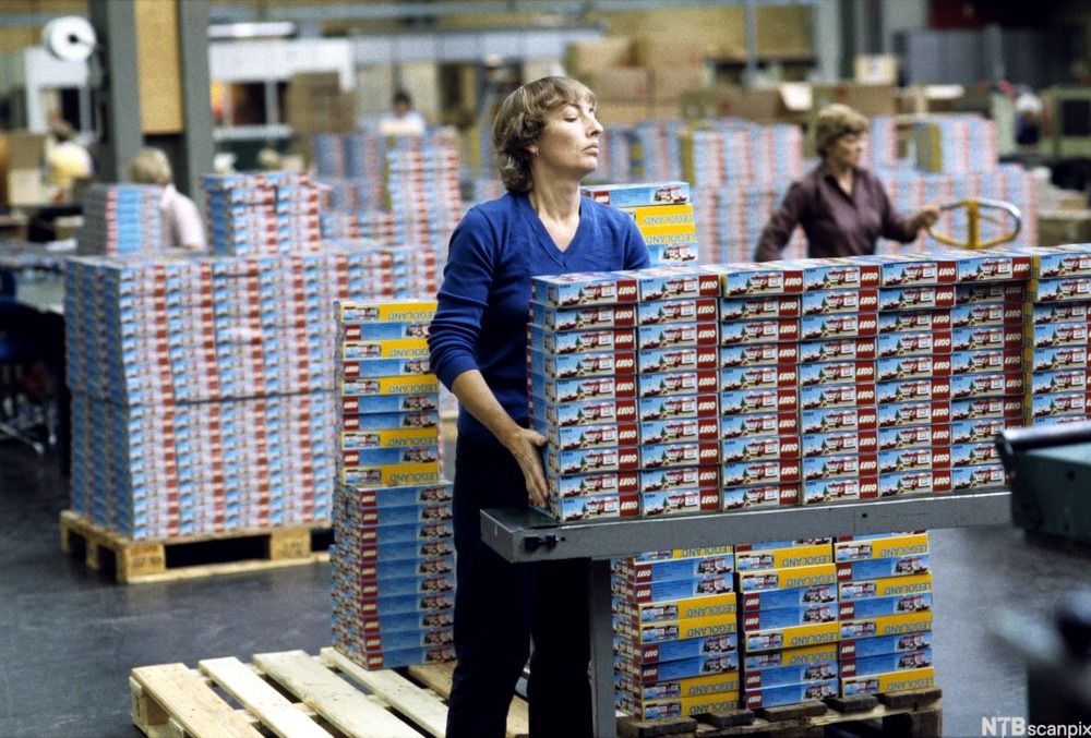 Legofabrikk: Ei kvinne plasserer fleire esker med lego som ligg på eit løftesystem. Det er fleire arbeidarar og stablar med legoesker i bakgrunnen. Foto.