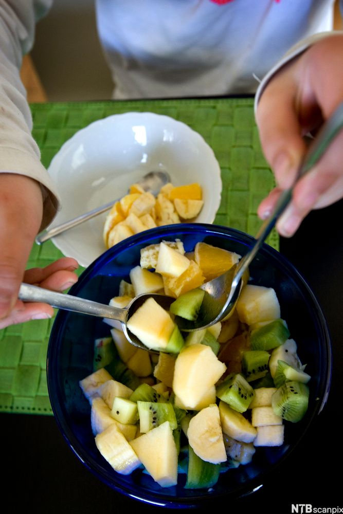 Person forsyner seg av fruktsalat med kiwi, banan, eple og appelsin. Foto.