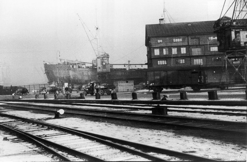 Et fraktskip ligger til havn. I forkanten er det jernbaneskinner. Svart-hvitt foto. 