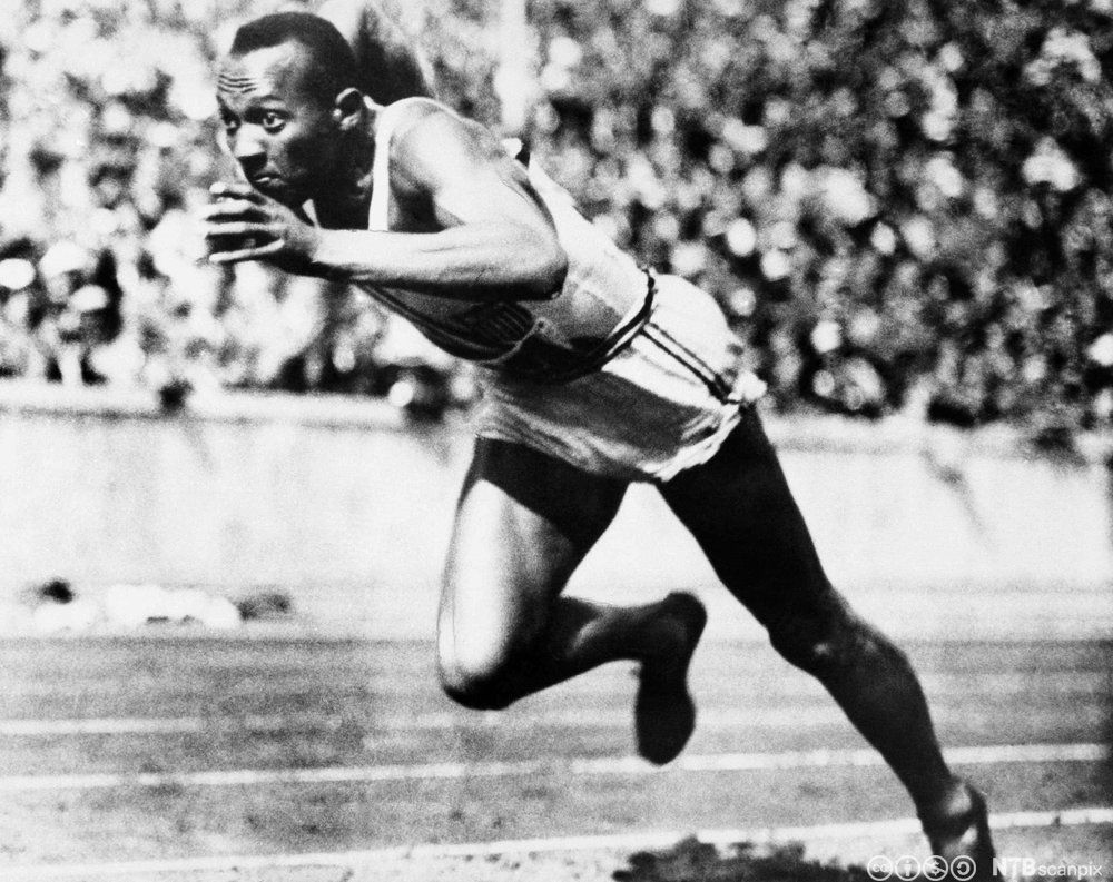 Svart mann i singlet og shorts løper framoverlent på en friidrettsbane. Fulle tribuner i bakgrunnen. Foto.