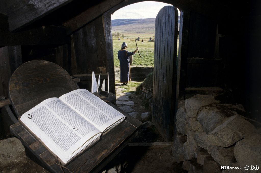 Utsikt fra underjordisk gang til islandsk landskap og mann med hjelm og øks. I forgrunnen ligger ei oppslått bok. Foto.