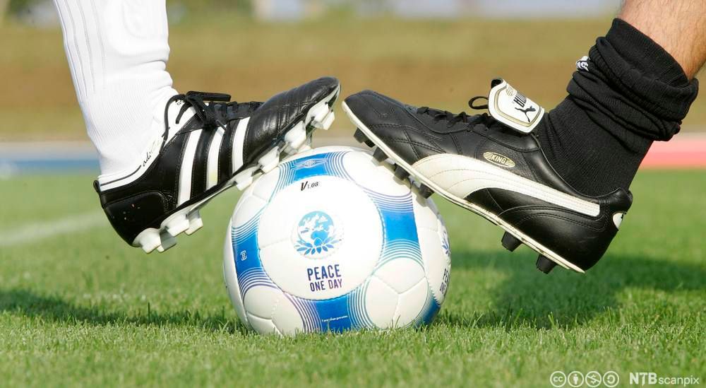 En fotball i midten, en fot i Adidas-sko og en fot i Puma-sko på hver si side av fotballen. Foto.