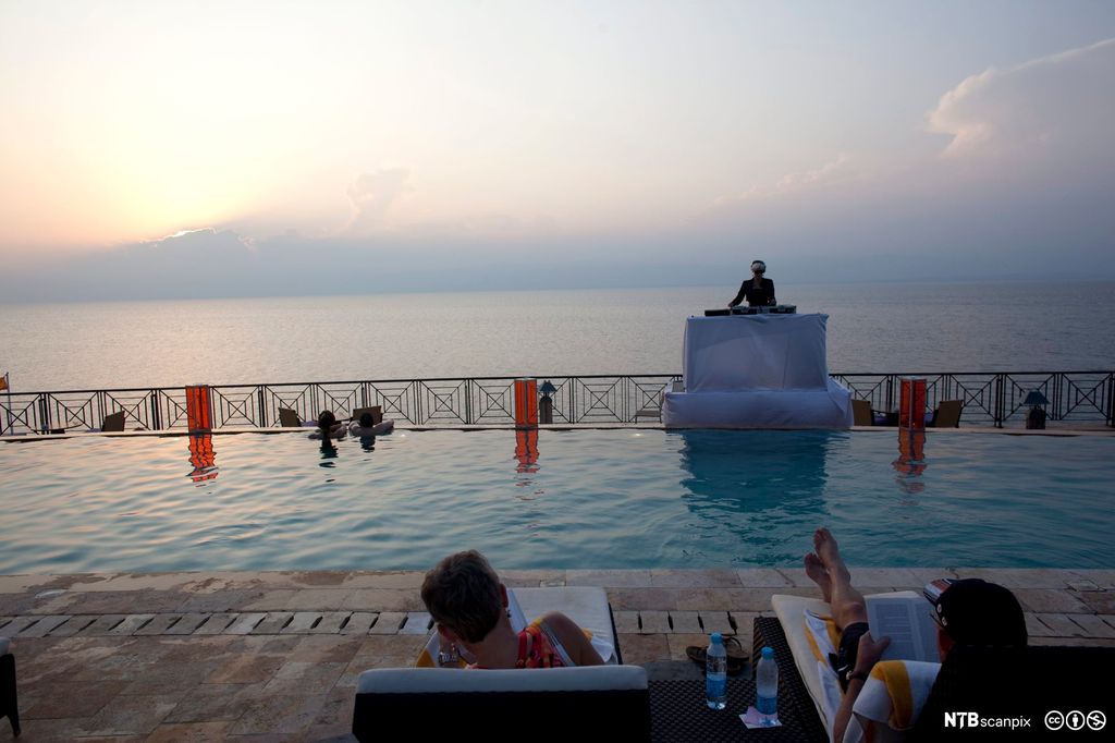 To personer ligger og leser på hver sin solseng ved ein bassengkant med utsikt mot havet og horisonten. Foto.