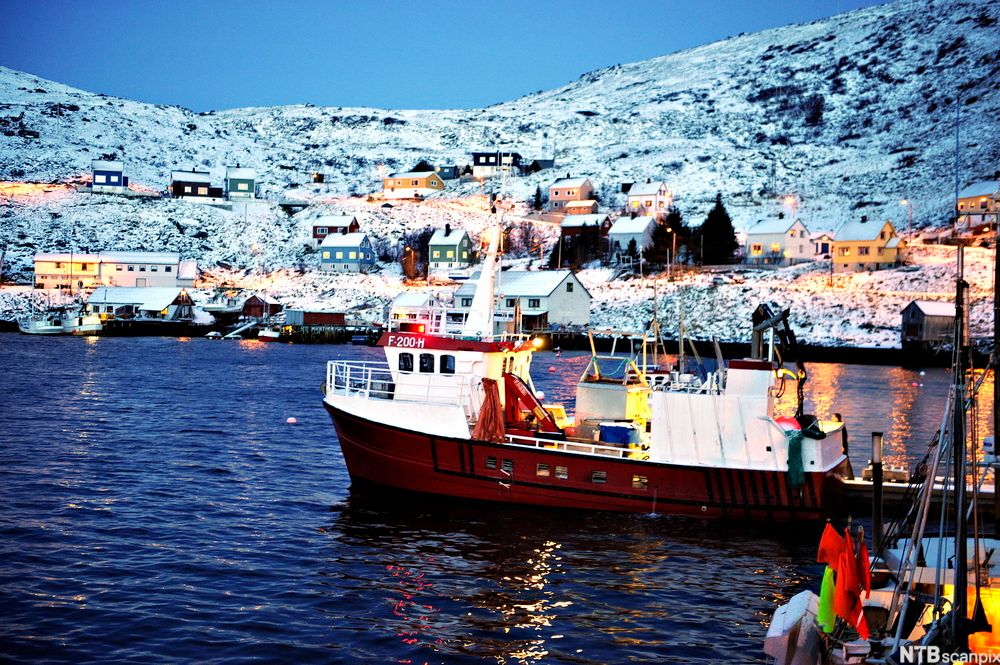 En fiskebåt har lagt fra kai. Den passerer ikke langt fra land. Ei snødekt åsside med spredt bebyggelse i bakgrunnen. Foto.
