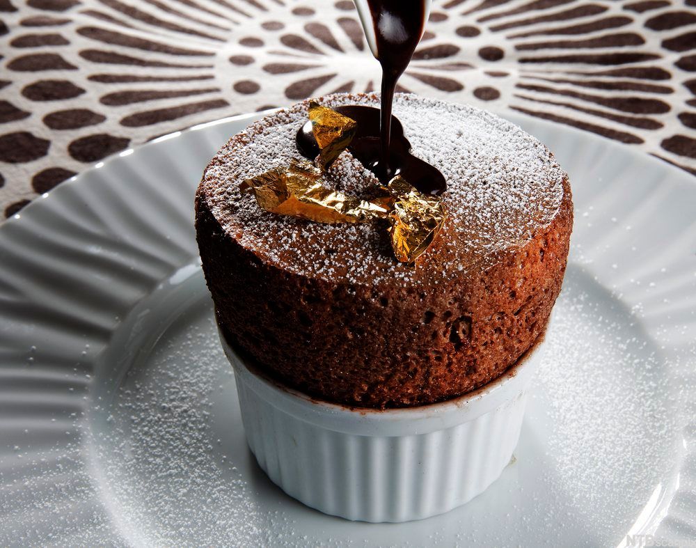 En ferdig stekt sjokoladesufflé i en hvit porsjonsskål, servert på en hvit tallerken. Suffléen er drysset med melis og pyntet med smeltet sjokolade og bladgull. Foto.