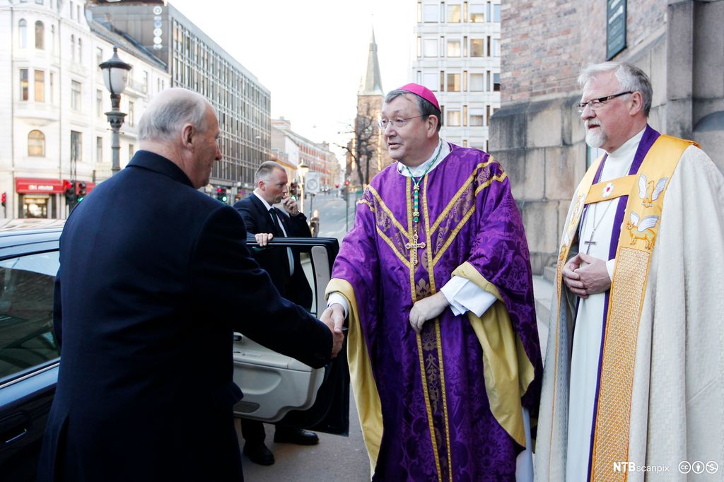To biskoper kledd i kjortel og messehagel hilser på en mann i frakk. Foto.