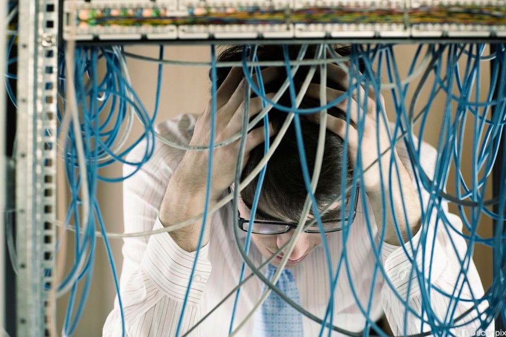 Mann sitter bak et virvar av kabler og ser ned mens han holder hodet i hendene. Foto. 