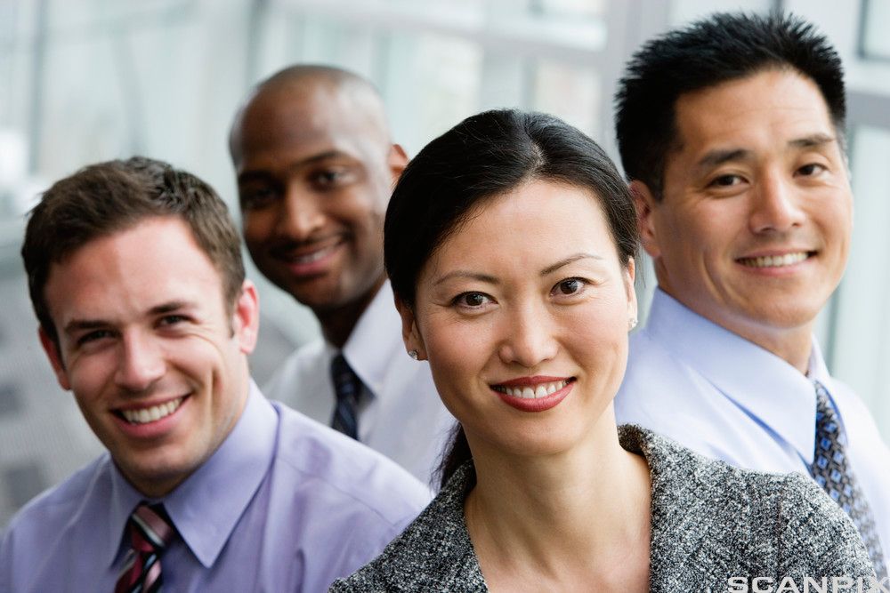 Fire smilende voksne personer i formelle antrekk som vender seg mot kamera. En kvinne foran og tre menn i bakgrunnen. Foto.