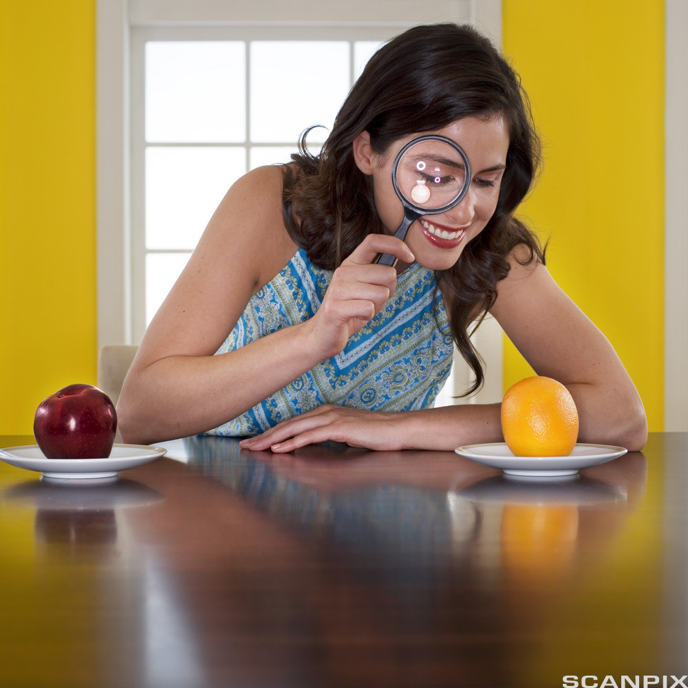 Kvinne undersøker ei appelsin som ligger på et tefat med et forstørrelsesglass. Foto.