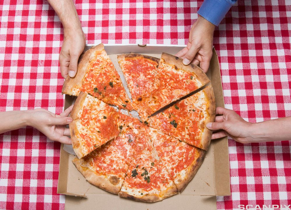 Flere personer tar hver sitt pizzastykke. Nærbilde av en stor, rund pizza og hender som griper etter stykkene. Foto.