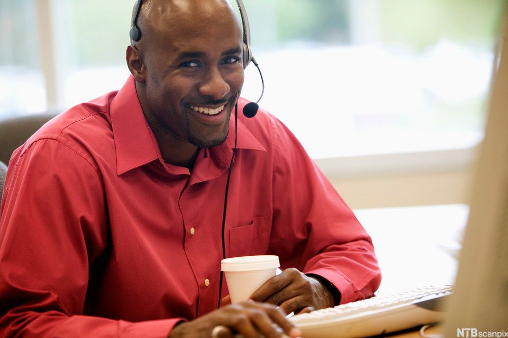 En mann sitter foran en datamaskin på et kontor. Han har på seg hodetelefoner og snakker inn i mikrofonen. Foto.