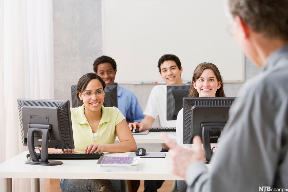 Elever ved datamaskiner som følger med på lærer som forklarer. Foto