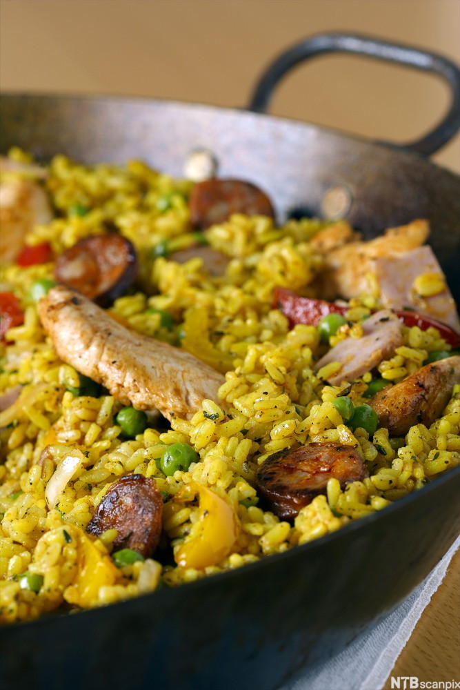 Paella i ei panne. Retten er fargerik med gul ris, grønne erter, rød paprika og biter av stekt kjøtt. Foto.
