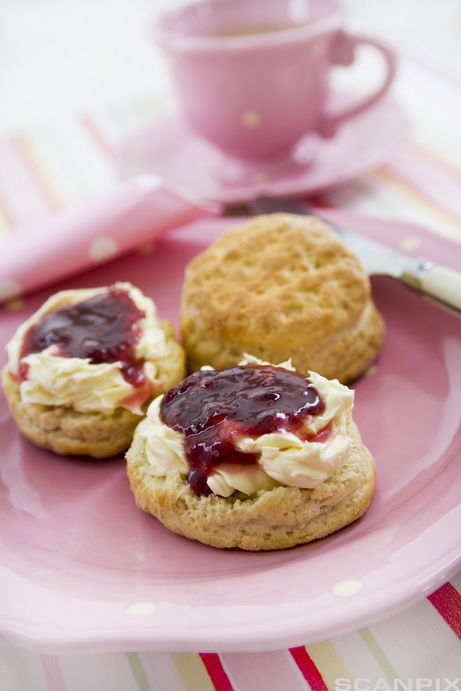 Tre scones på en rosa tallerken, to har syltetøy på seg. En tekopp står ved siden. Foto.
