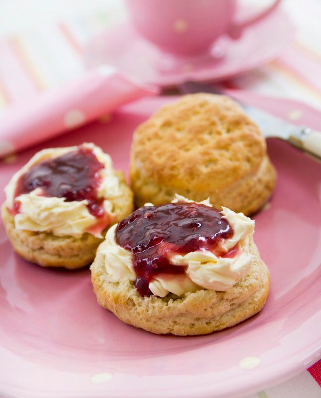 Tre scones på en rosa tallerken, to har syltetøy på seg. En tekopp står ved siden. Foto.