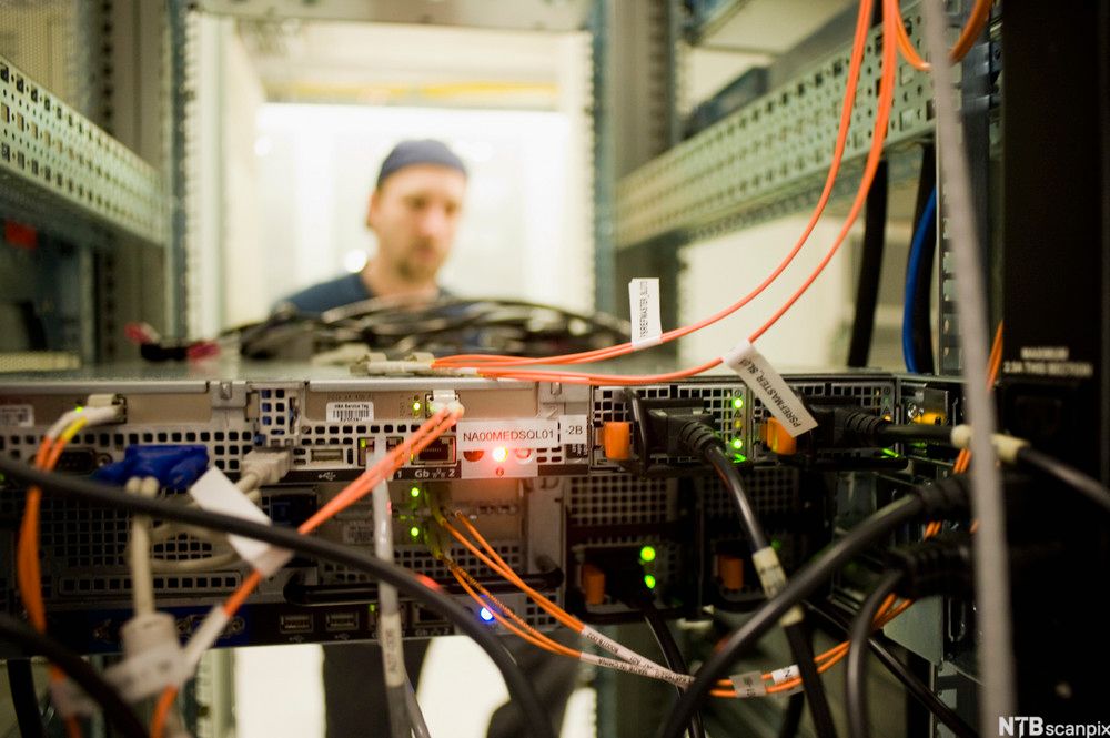 Baksiden av en server med mange kabler, i bakgrunnen ser en mann inn på serveren. Foto.