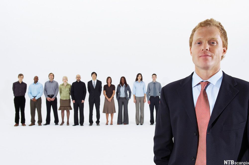 En person står i forgrunnen nær kamera. I bakgrunnen står ti personer på rekke, og disse er litt ute av fokus. Foto.