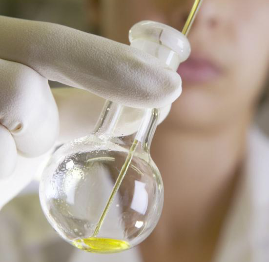 Ein person med kvite hanskar held opp ein kolbe med pipette og oljeekstrakt. Foto.