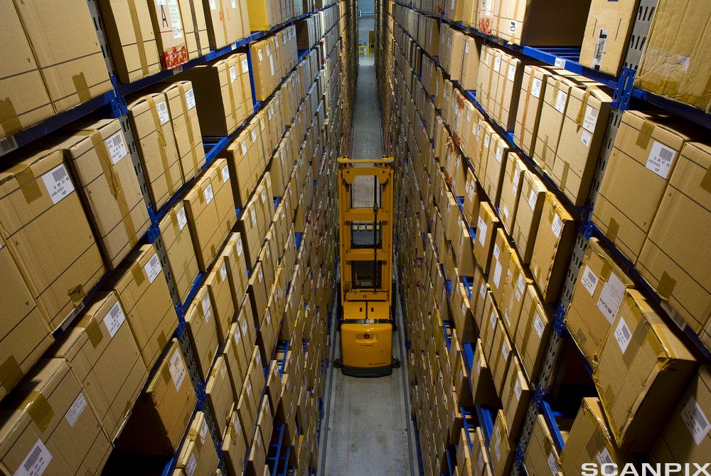 Store rader med hyller med pappesker. Gaffeltruck står mellom radene. Foto.