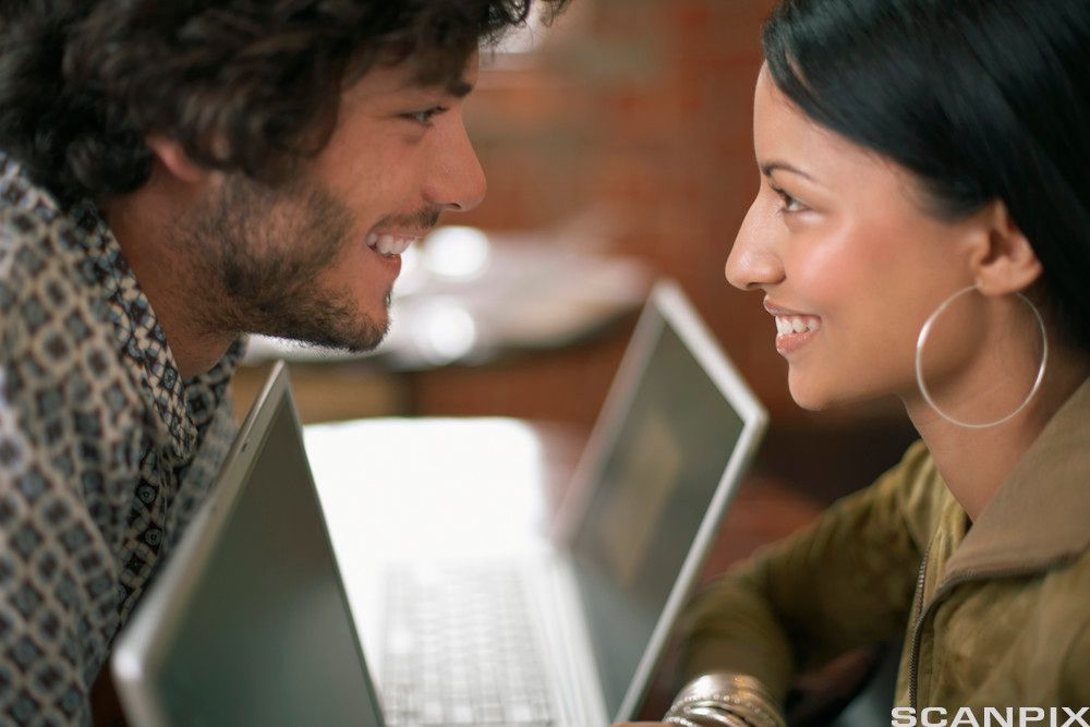 En ung mann og en ung kvinne smiler til hverandre. Foran seg har de hver sin bærbare PC. Foto.