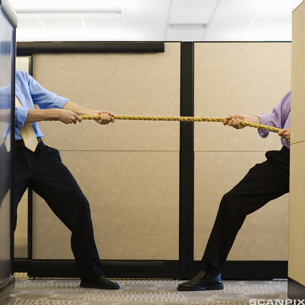 To menn i skjorte og slips står delvis skjult bak hver sin skillevegg på en arbeidsplass og drar i hver sin ende av et tau. Foto.