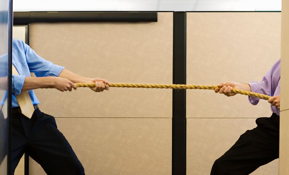 To menn i skjorte og slips står delvis skjult bak hver sin skillevegg på en arbeidsplass og drar i hver sin ende av et tau. Foto.