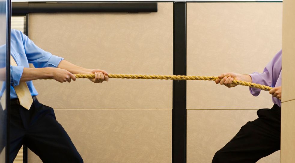 To menn i skjorte og slips står delvis skjult bak hver sin skillevegg på en arbeidsplass og drar i hver sin ende av et tau. Foto.