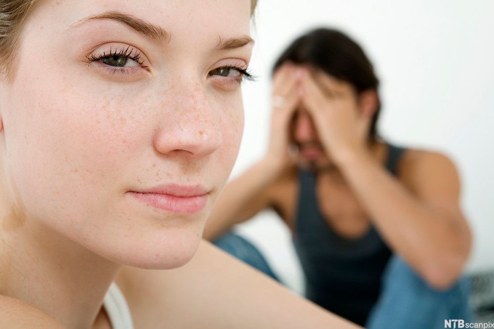 Kvinne med skulende blikk i forgrunnen og mann med hodet i hendene i bakgrunnen. Foto.