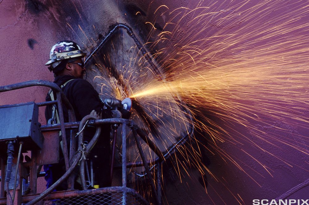 Person som bruker vinkelsliper på en stor stålkonstruksjon. Personen bruker verneutstyr, og slipinga gir mye gnistsprut. Foto.